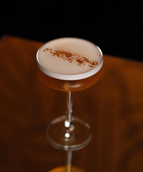 Cocktail Amaretto sour servi dans une coupe, coiffé d'une mousse onctueuse et saupoudré de cannelle ou d'épices, sur fond sombre.